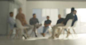 A group of men sitting in a circle in a well lit room. Image is out of focus.