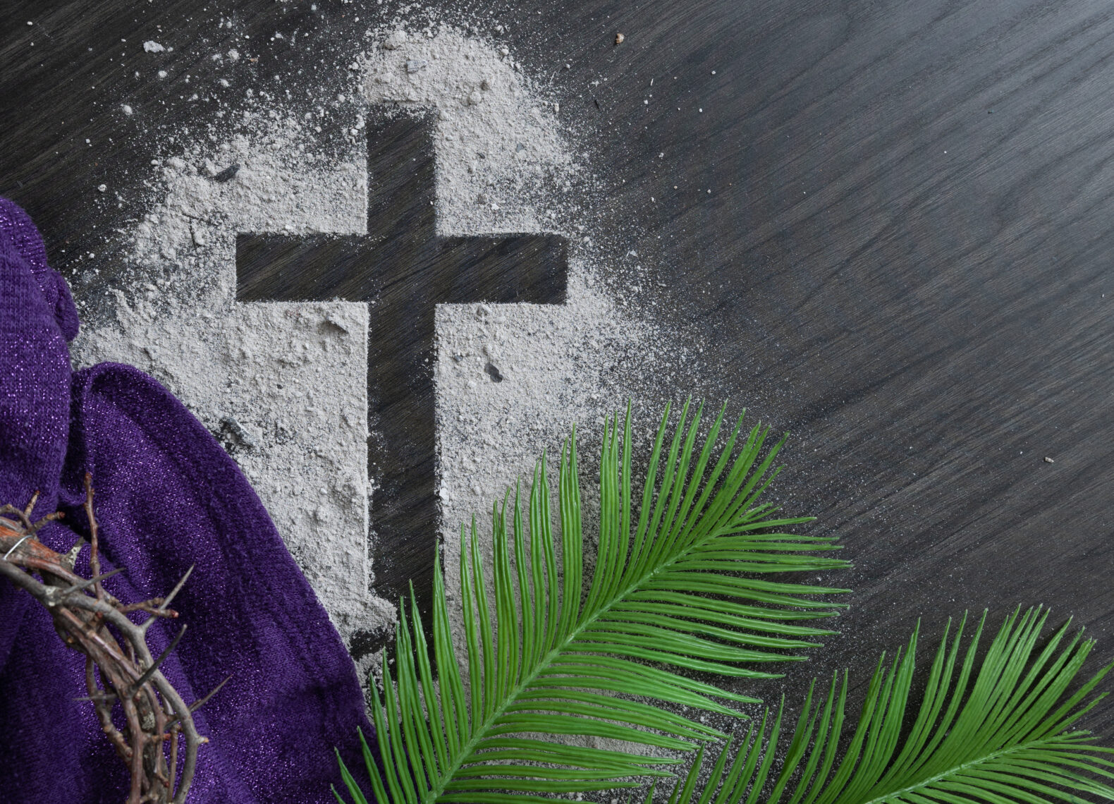 Christian lent background with cross of ashes, palm fronds, purple cloth and partial crown of thorns on a dark wood background with copy space